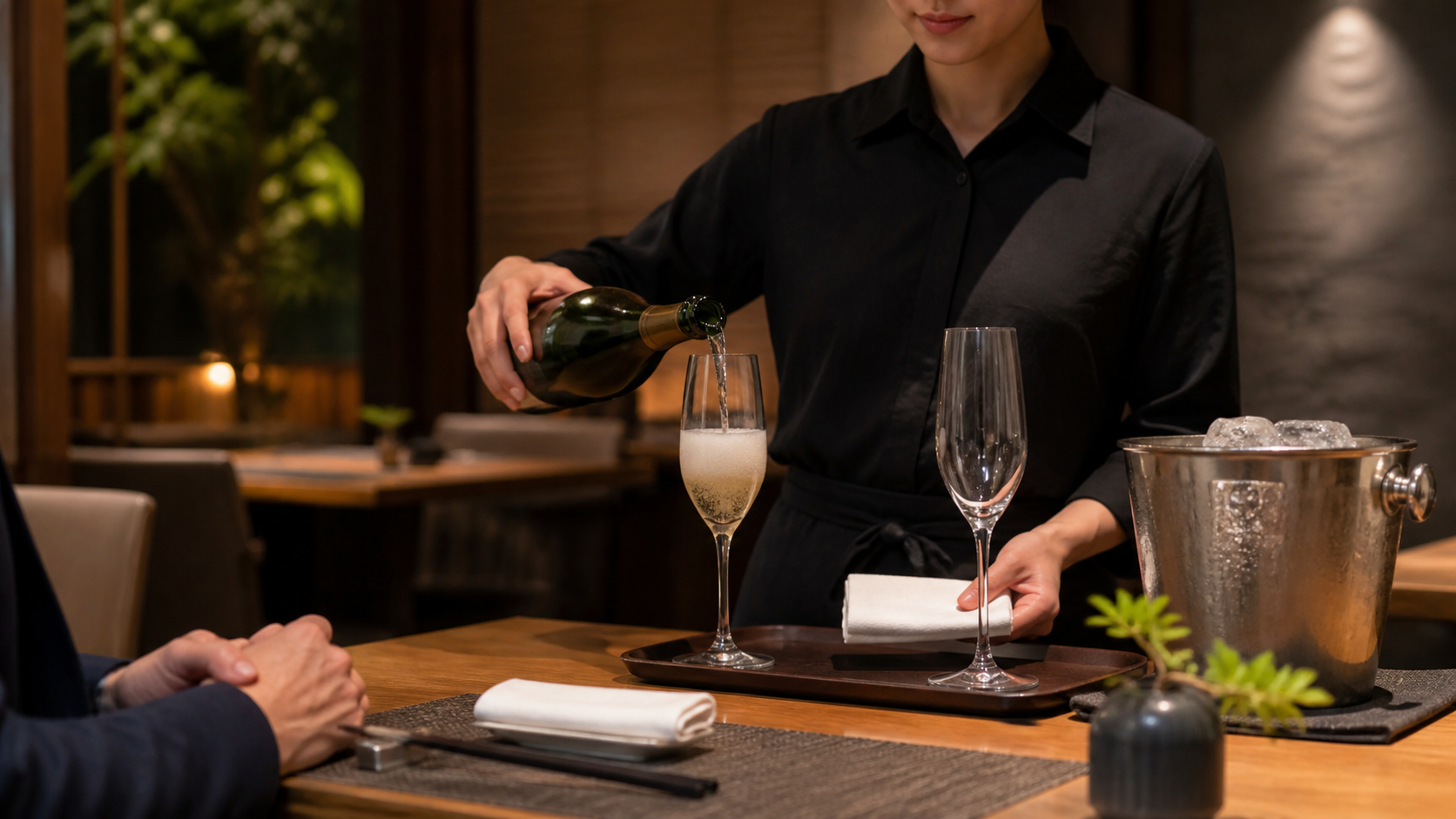 飲食店のスタッフが、泡が落ち着くのを待つため最初に少量を注いだスパークリングワインのグラスと、次に注ぐ空のグラスをテーブル脇で用意している場面