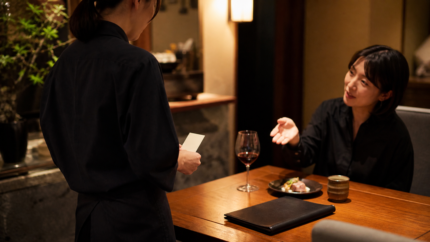 飲食店の客席で、スタッフが短いカードを確認しながら赤ワインを飲むお客様の会話を聞いている場面